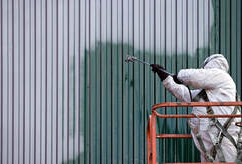 Professional painter in Magog wearing full protective equipment on a gondola. He paints the siding of a metal building.